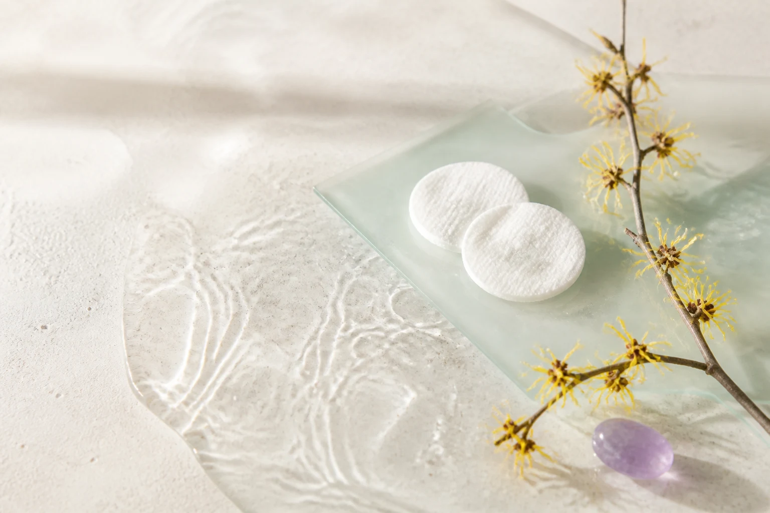 Dischetti di cotone, acqua, vetro celadon e fiori di amamelide in una scena luminosa
