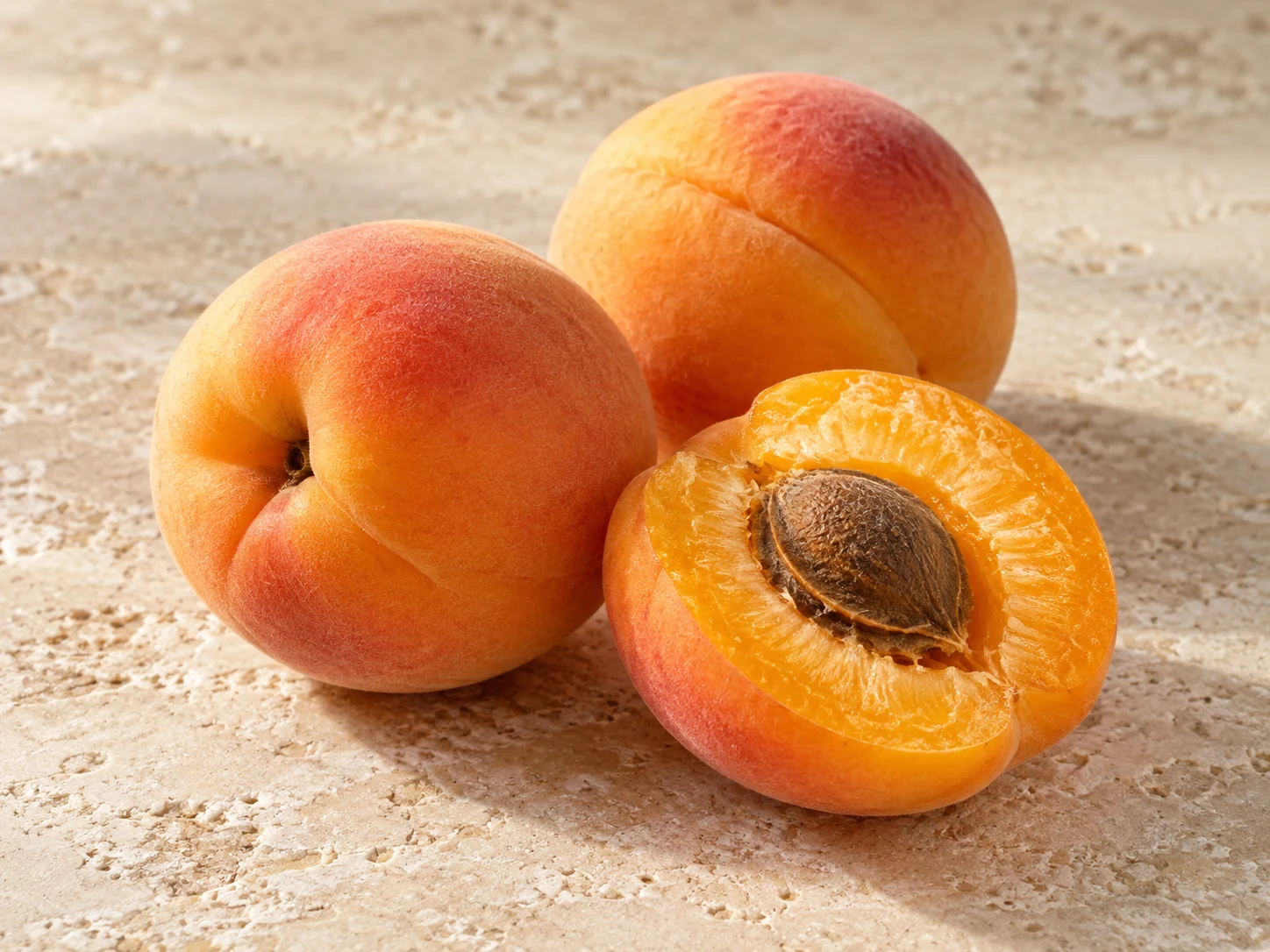 Due albicocche dorate e una metà con polpa arancione e nocciolo su pietra chiara.